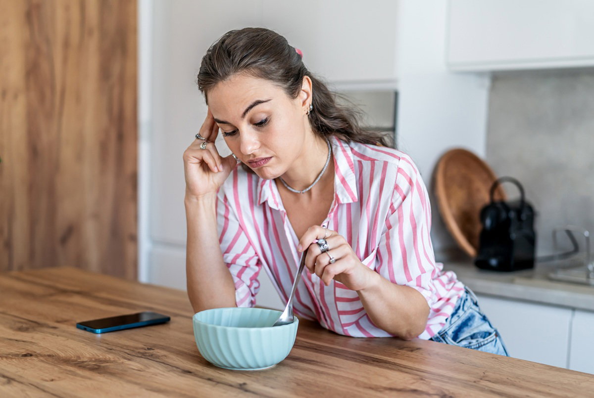 kobieta w wieku 30 plus w koszuli w paski miesza łyżką w miseczce stojąc w kuchni z obrażoną miną