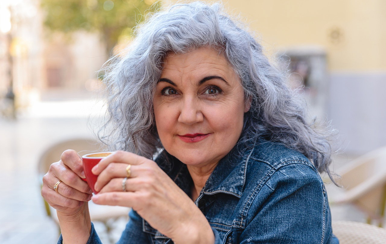 kobieta w wieku 60 plus z siwymi włosami w jeansowej koszuli siedzi na dworze i pije kawę zadowolona z siebie