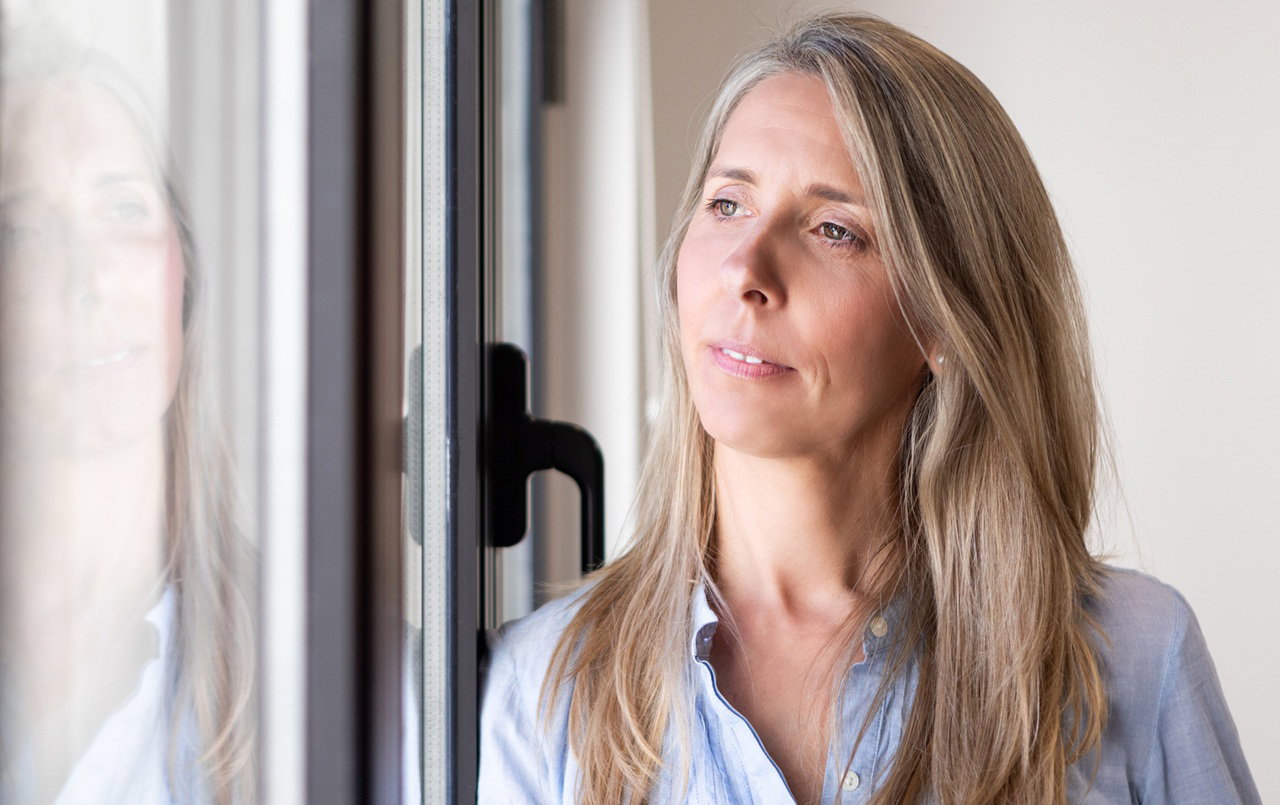 kobieta w wieku 30 plus w błękitnej koszuli patrzy przez okno zamyślona