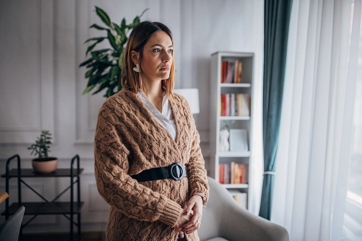 kobieta w  wieku 40 plus w beżowym swetrze patrzy smutna przez okno