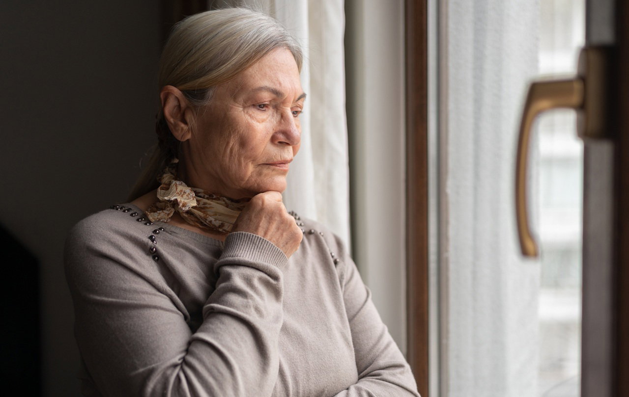kobieta w wieku 65 plus z siwymi włosami i apaszka na szyi w szarym swetrze stoi i patrzy smutno przez okno