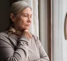 kobieta w wieku 65 plus z siwymi włosami i apaszka na szyi w szarym swetrze stoi i patrzy smutno przez okno