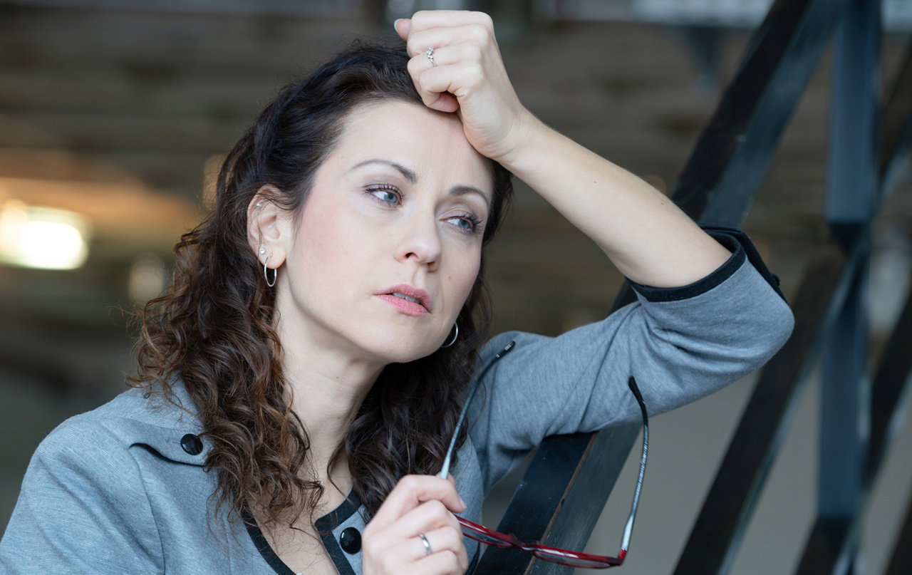 kobieta w wieku 30 plus w szarym żakiecie z długimi kręconymi włosami stoi zamyślona z okularami w ręku