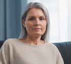 kobieta w wieku 40 plus siwe włosy w beżowym swetrze siedzi na kanapie smutna