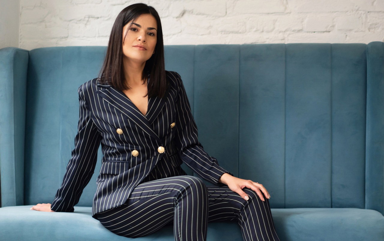 kobieta w wieku 30 plus ciemne włosy w dopasowanym garniturze siedzi na sofie z poważną miną