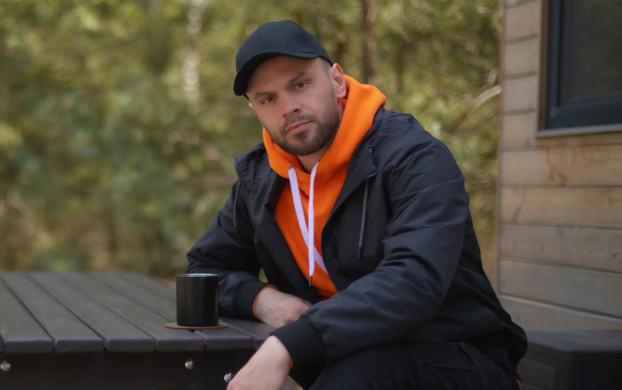 mężczyzna w wieku 30 plus w czapce z daszkiem siedzi przy drewnianej ławie z kubkiem herbaty zamyślony