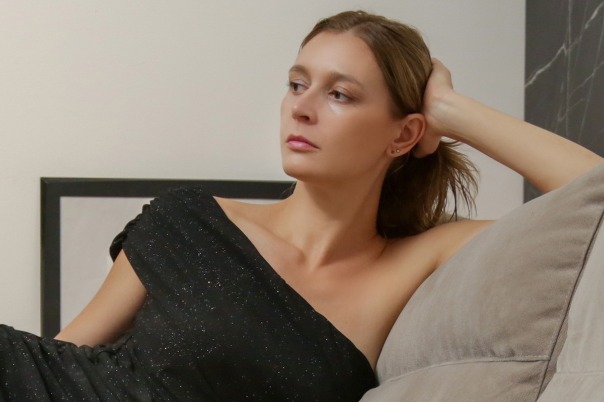 kobieta w wieku 30 plus w ciemnej mieniącej się sukni siedzi zamyślona i smutna na sofie