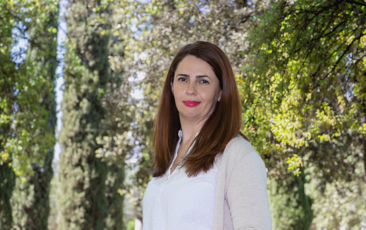 kobieta w wieku 35 plus w białej bluzce i jasnym sweterku stoi na zewnątrz na tle drzew z kpiącą miną