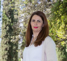 kobieta w wieku 35 plus w białej bluzce i jasnym sweterku stoi na zewnątrz na tle drzew z kpiącą miną