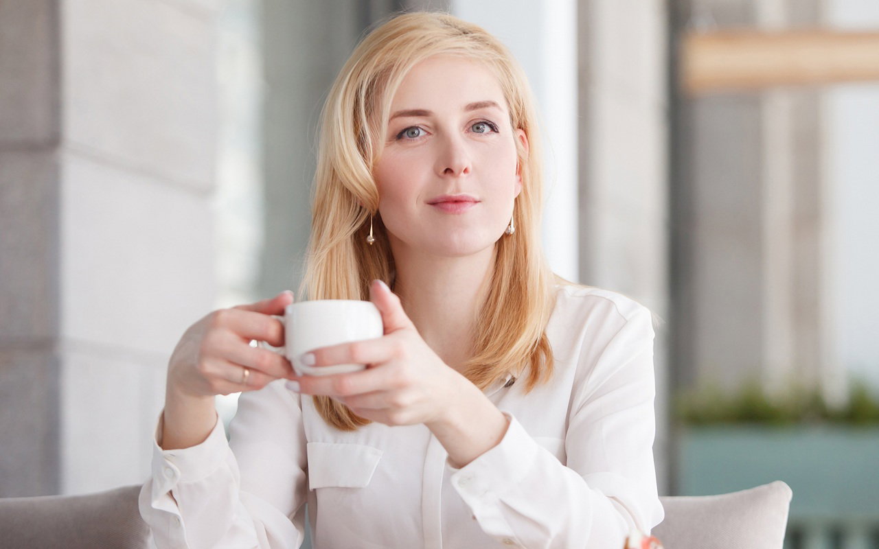 kobieta w wieku 30 plus w białej bluzce blondynka siedzi w restauracji z kawą i lekko się uśmiecha