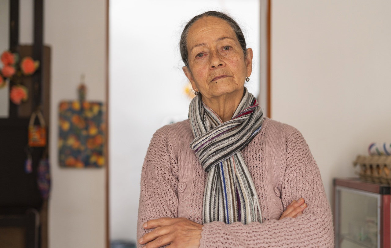 kobieta w wieku 70 plus z szalikiem w różowym sweterku stoi na tle kuchni z poważną miną