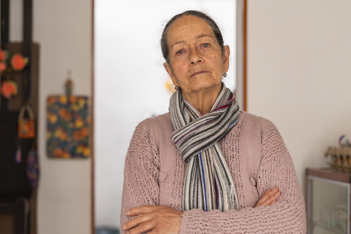 kobieta w wieku 70 plus z szalikiem w różowym sweterku stoi na tle kuchni z poważną miną
