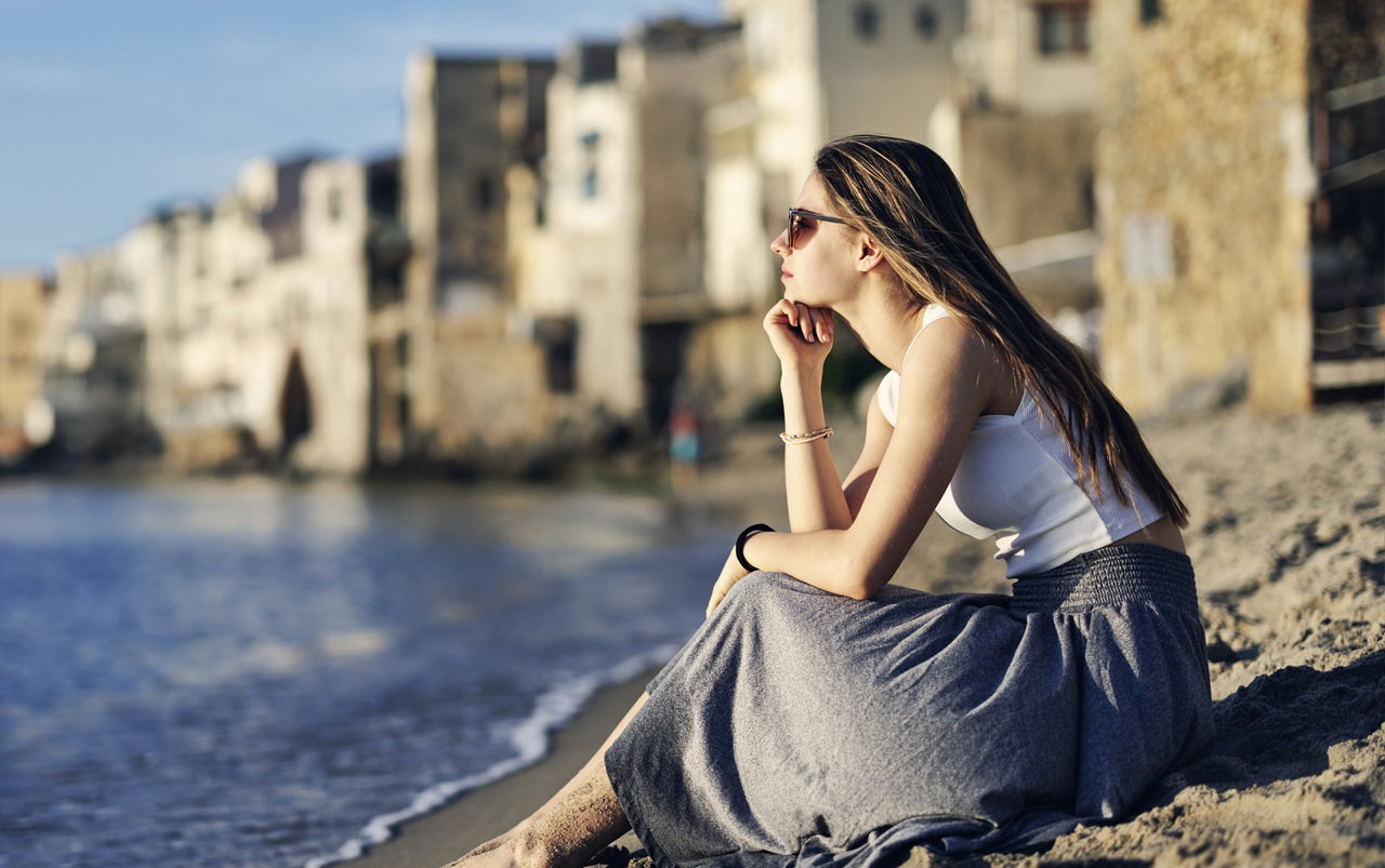 kobieta w wieku 25 plus siedzi na plaży w miasteczku Cefalu na Sycylii i patrzy w dal zamyślona