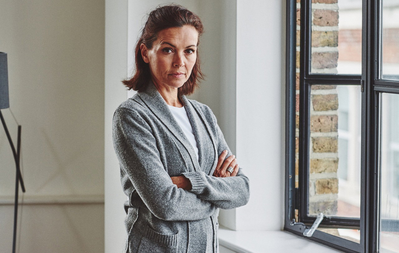 kobieta w wieku 40 plus w szarym swetrze stoi przy oknie poirytowana