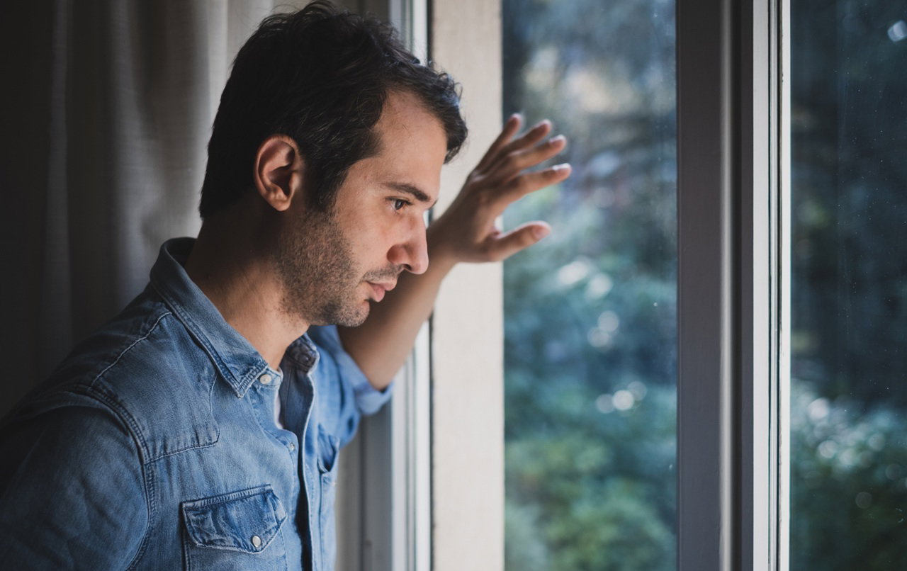 mężczyzna w wieku 35 plus w jeansowej koszuli stoi przy oknie zamyślony i smutny