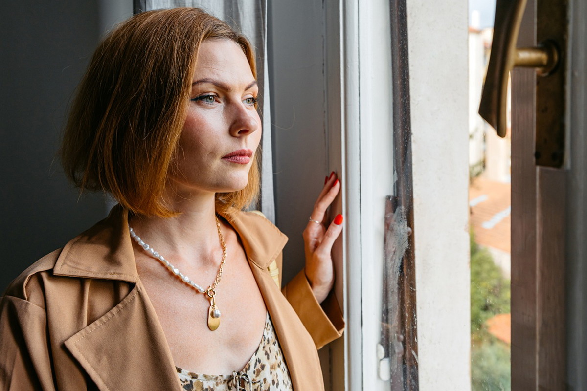 kobieta w wieku 30 plus rude włosy stoi w płaszczu przy oknie zamyślona i smutna