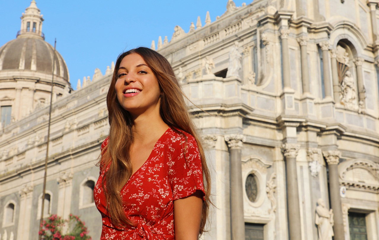 kobieta w  wieku 30 plus w czerwonej sukience w Katanii we Włoszech stoi na tle katedry i się uśmiecha