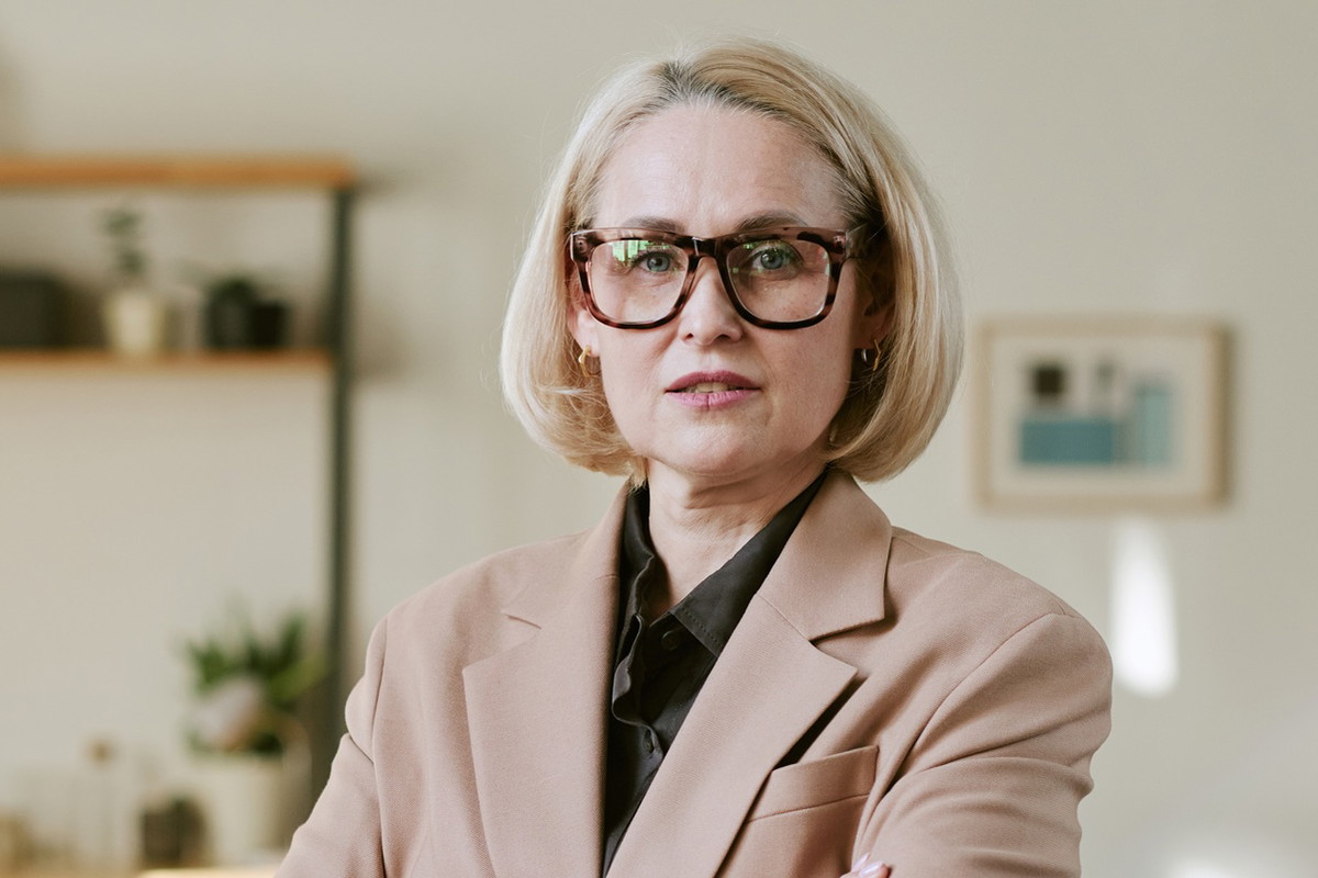 kobieta w wieku 40 plus blondynka w okularach stoi na tle pokoju z poważną miną