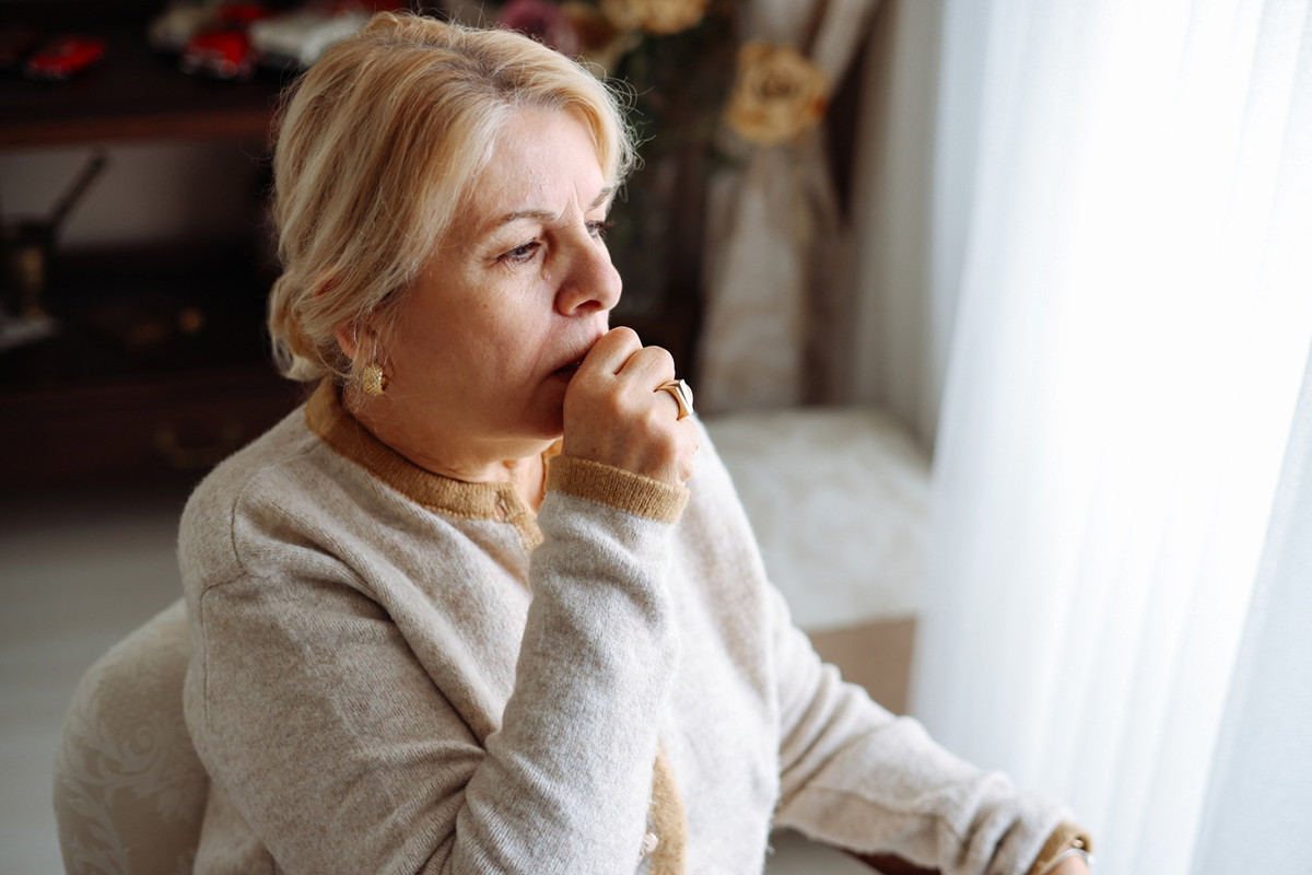 kobieta w wieku 65 plus blond włosy siedzi przy oknie w swoim pokoju zamyślona i smutna