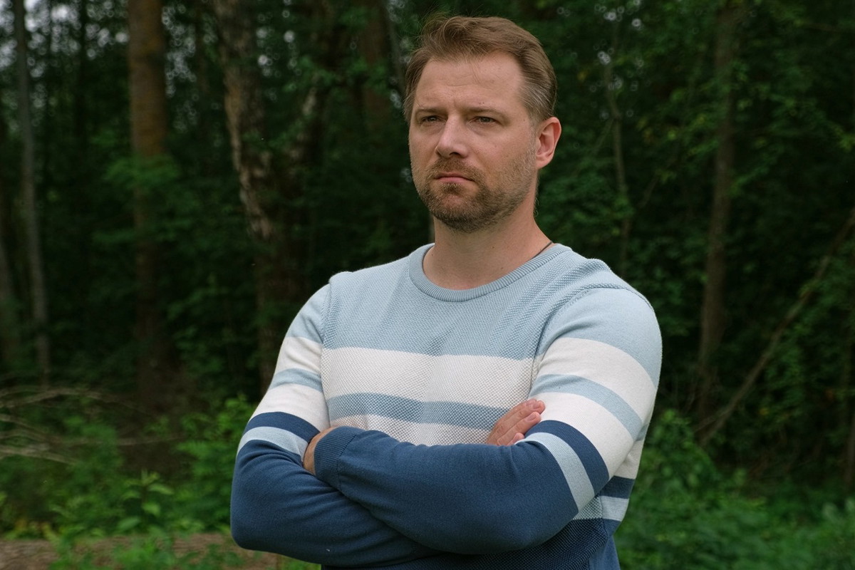 mężczyzna w wieku 30 plus w niebieskim sweterku w pasy stoi na tle drzew nachmurzony