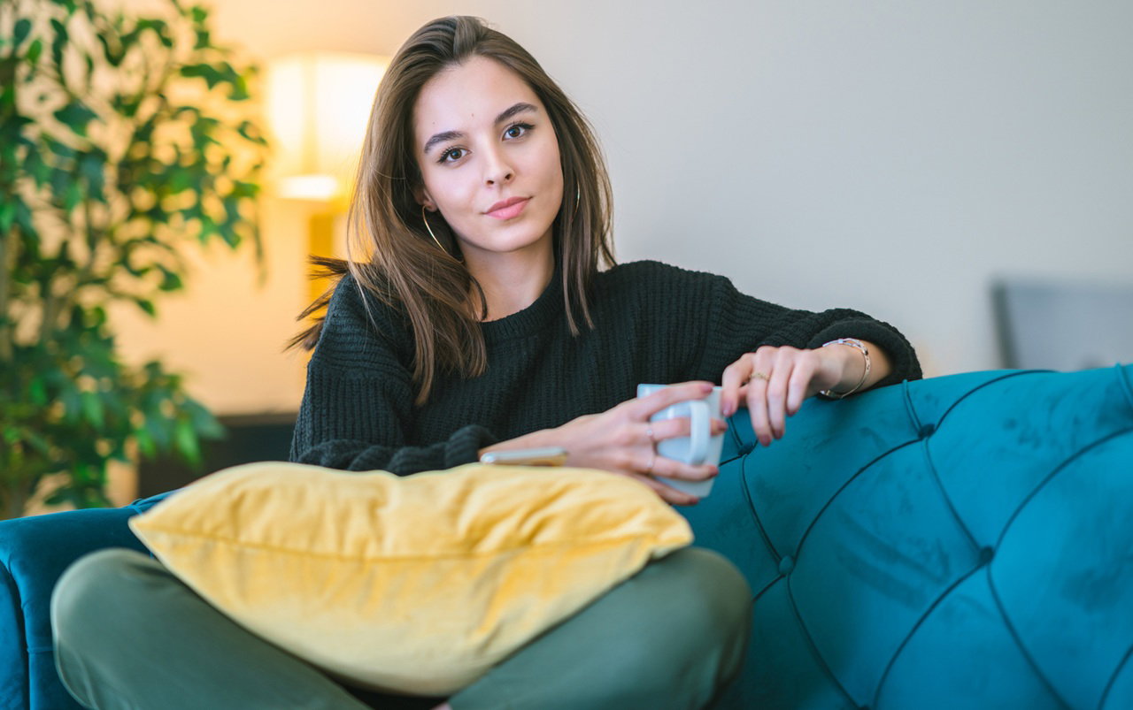 kobieta w wieku 25 plus siedzi na niebieskiej sofie w kubkiem w ręku z lekko kpiącą miną