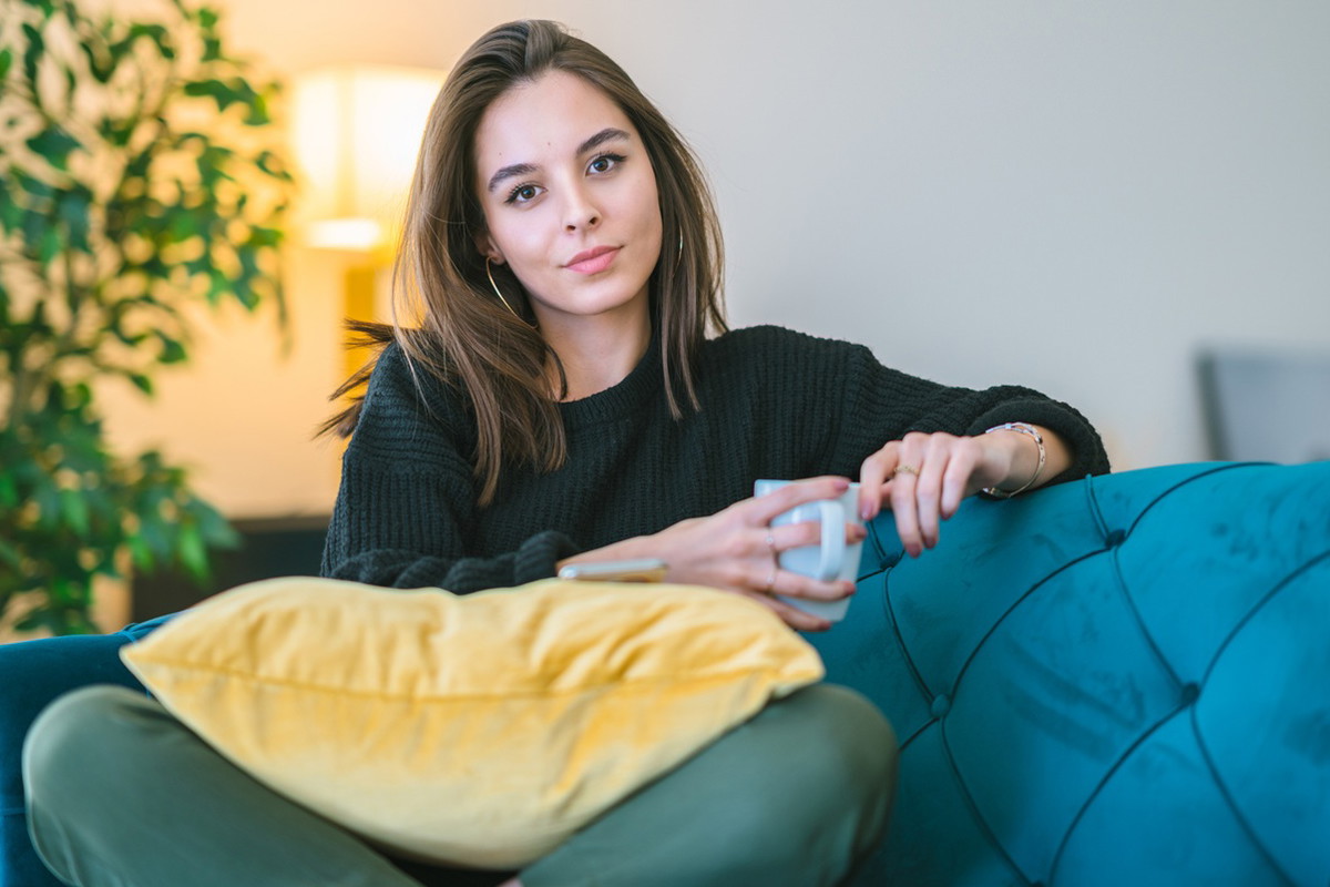 kobieta w wieku 25 plus siedzi na niebieskiej sofie w kubkiem w ręku z lekko kpiącą miną