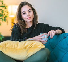 kobieta w wieku 25 plus siedzi na niebieskiej sofie w kubkiem w ręku z lekko kpiącą miną