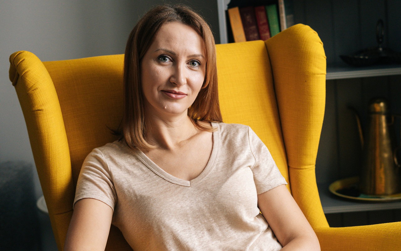 kobieta w wieku 30 plus siedzi w żółtym fotelu uszaku i lekko się uśmiecha z przekąsem
