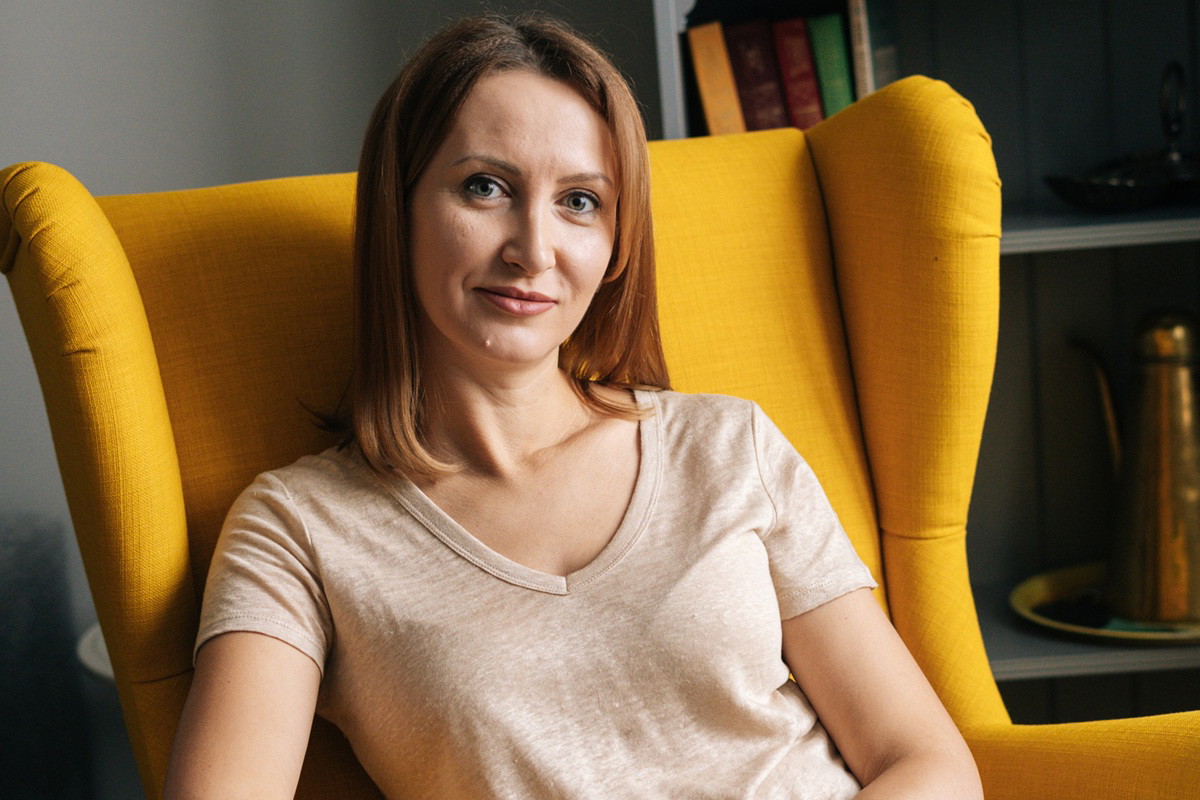 kobieta w wieku 30 plus siedzi w żółtym fotelu uszaku i lekko się uśmiecha z przekąsem