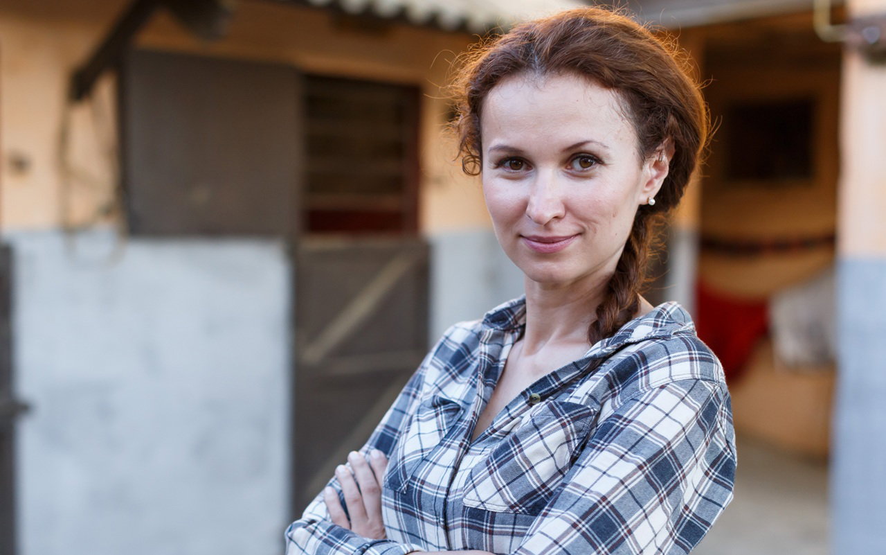 kobieta w wieku 30 plus w kraciastej koszuli stoi na tle stajni i uśmiecha się z przekąsem