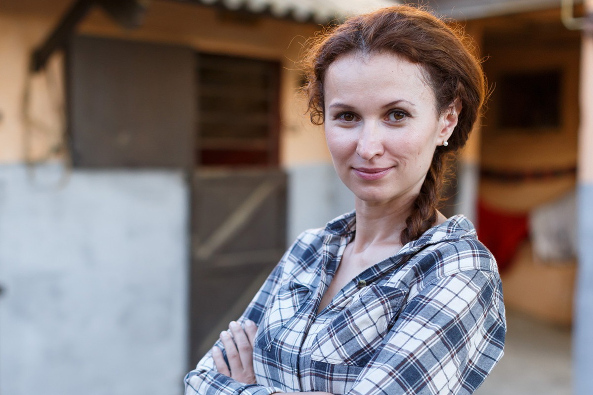 kobieta w wieku 30 plus w kraciastej koszuli stoi na tle stajni i uśmiecha się z przekąsem