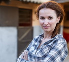 kobieta w wieku 30 plus w kraciastej koszuli stoi na tle stajni i uśmiecha się z przekąsem