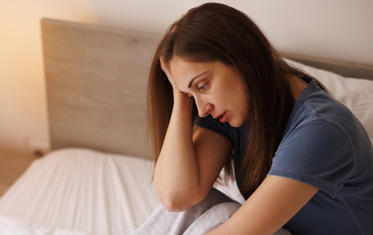 kobieta w wieku 35 plus ciemne włosy siedzi na łóżku w piżamie załamana