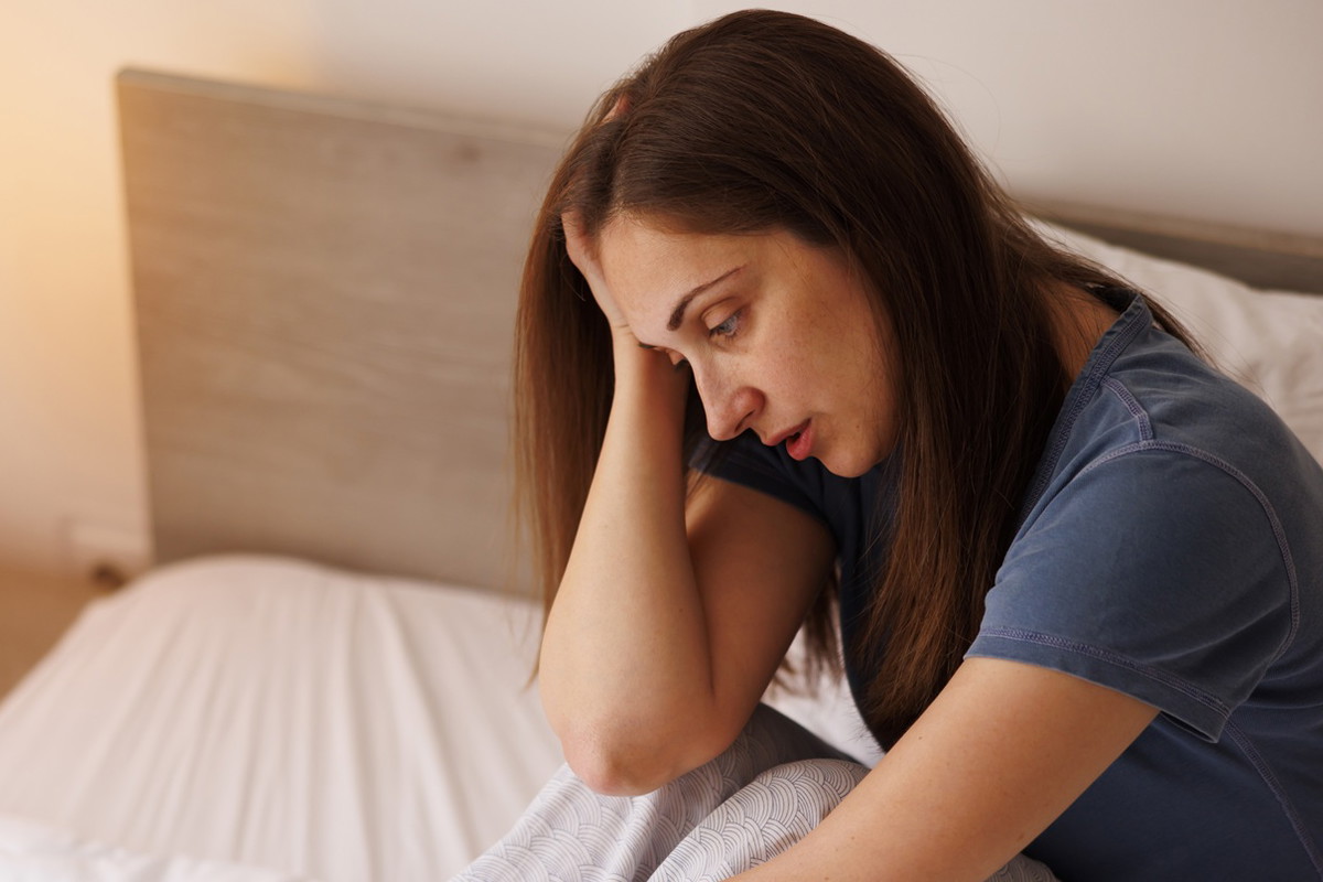 kobieta w wieku 35 plus ciemne włosy siedzi na łóżku w piżamie załamana