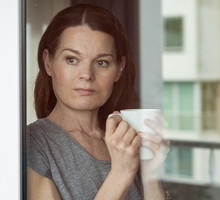 kobieta w wieku 40 plus patrzy przez okno zamyślona z kubkiem w ręku
