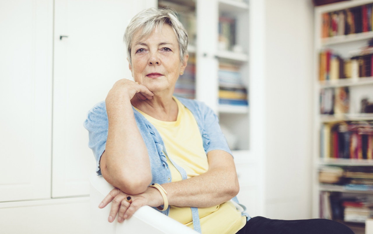 kobieta w wieku 55 plus siedzi na tle salonu z książkami zamyślona i smutna