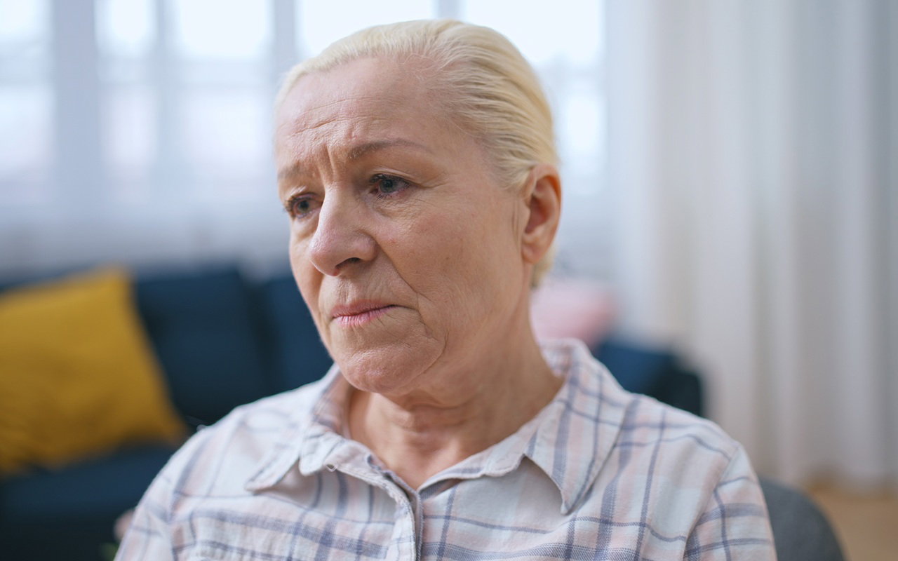 kobieta w wieku 65 plus blond włosy siedzi na kanapie  w salonie załamana i smutna