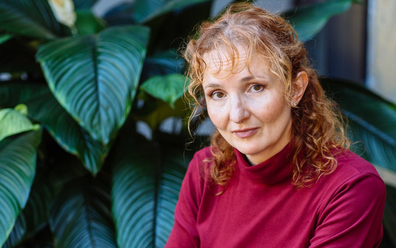 kobieta w wieku 45 plus z rudymi włosami w czerwonej bluzce siedzi na tle zielonej rośliny załamana