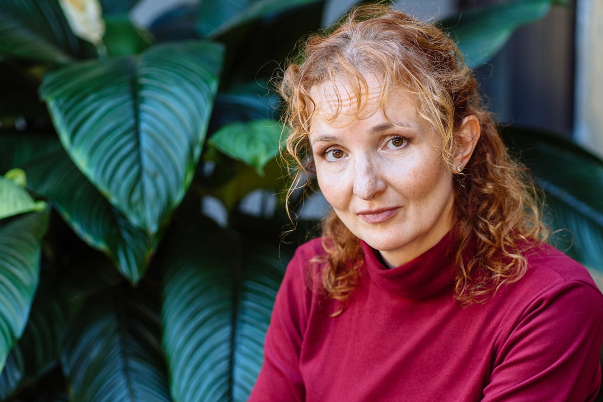 kobieta w wieku 45 plus z rudymi włosami w czerwonej bluzce siedzi na tle zielonej rośliny załamana
