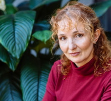 kobieta w wieku 45 plus z rudymi włosami w czerwonej bluzce siedzi na tle zielonej rośliny załamana