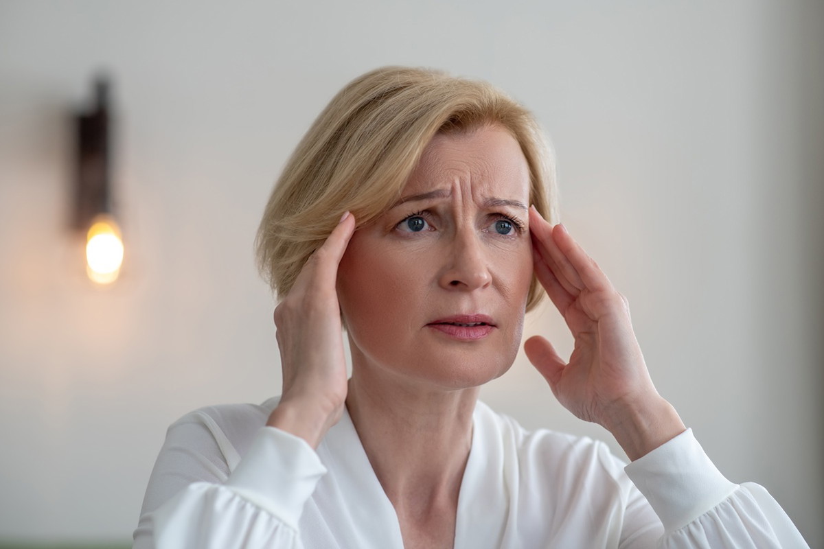 kobieta w wieku 50 plus blondynka w białej bluzce stoi zmarwiona i zrozpaczona w salonie
