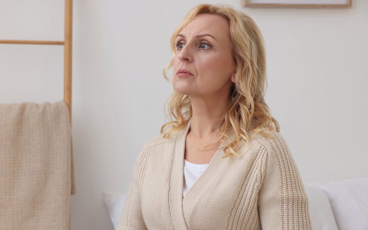 kobieta w wieku 55 plus blondynka w beżowym swetrze siedzi w pokoju lekko przerażona
