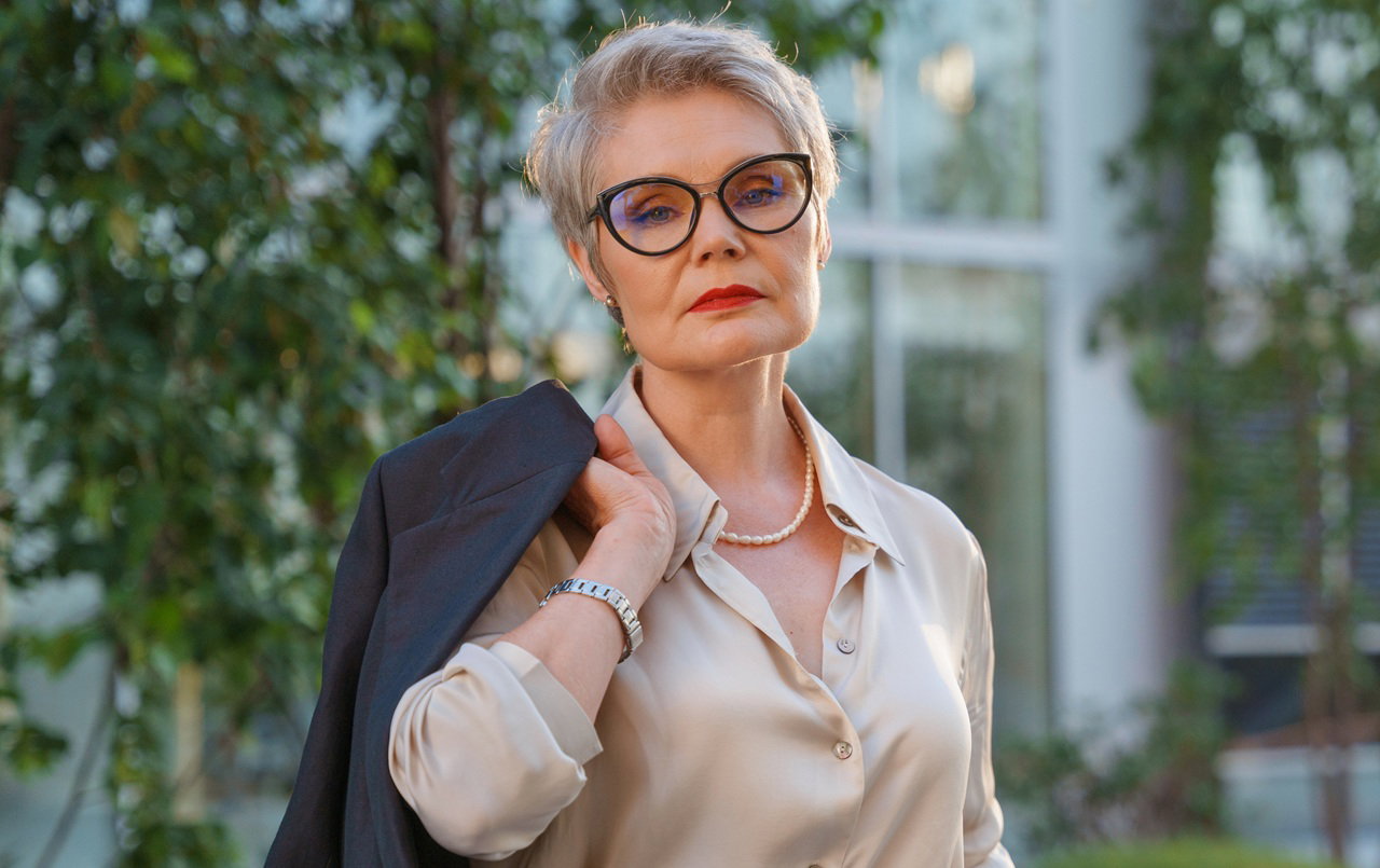 kobieta w wieku 55 plus w okularach elegancko ubrana stoi na tle budynku i zieleniu zrezygnowana