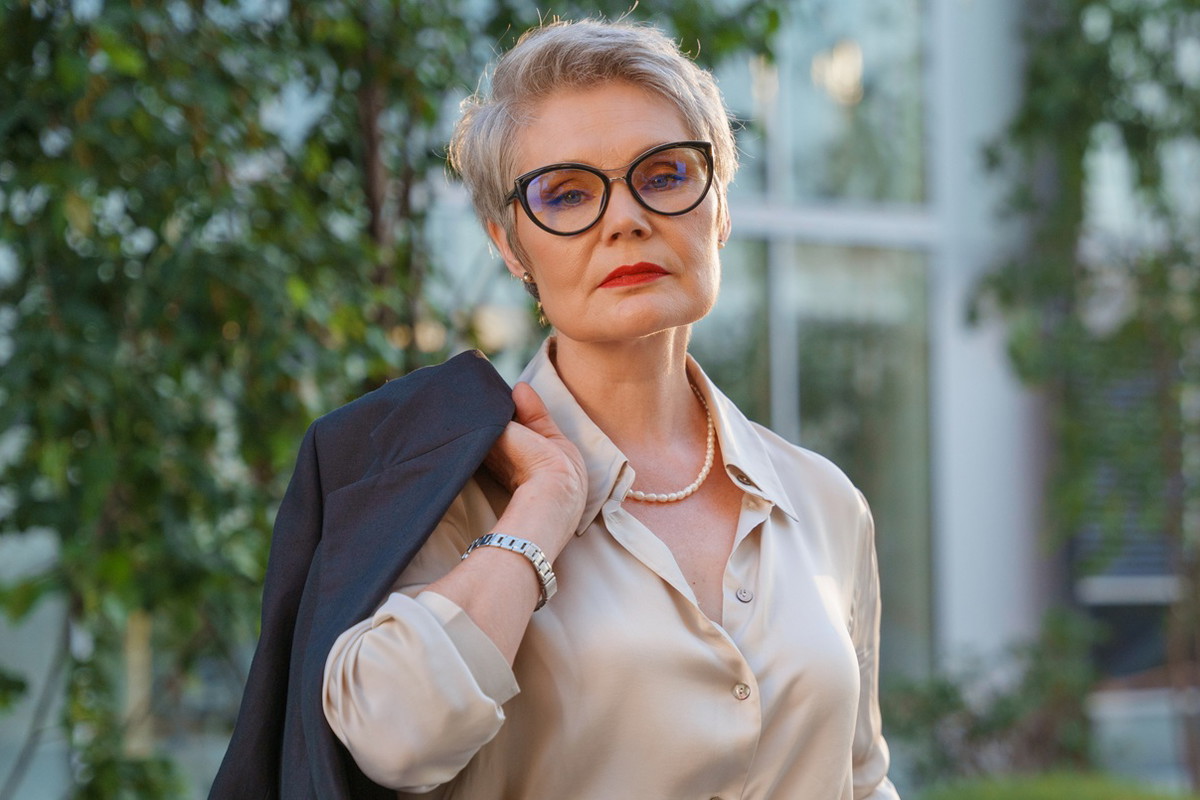 kobieta w wieku 55 plus w okularach elegancko ubrana stoi na tle budynku i zieleniu zrezygnowana