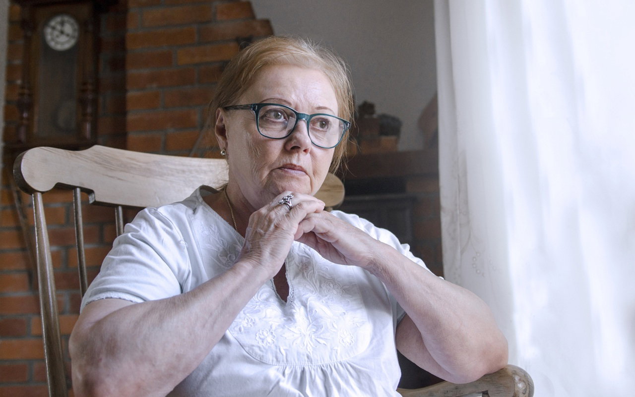 kobieta w wieku 60 plus w okularach siedzi na krześle w jasnej podomce zamyślona i smutna