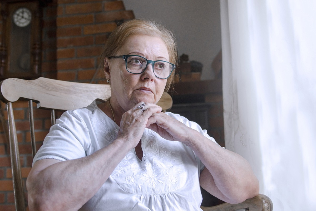 kobieta w wieku 60 plus w okularach siedzi na krześle w jasnej podomce zamyślona i smutna