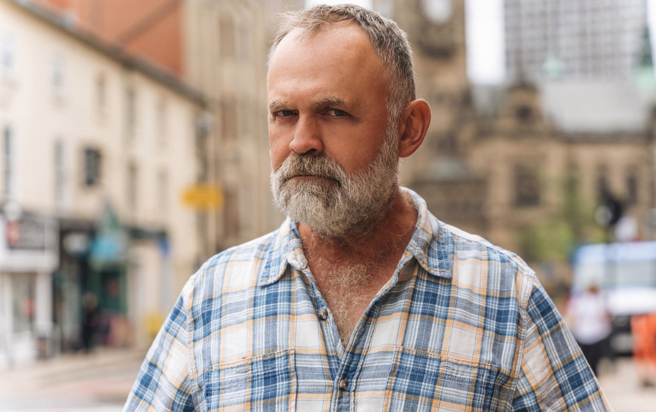 mężczyzna w wieku 50 plus w kraciastej koszuli stoi na tle miasta z poważną miną