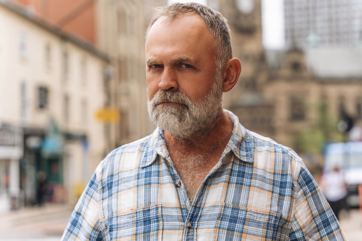 mężczyzna w wieku 50 plus w kraciastej koszuli stoi na tle miasta z poważną miną