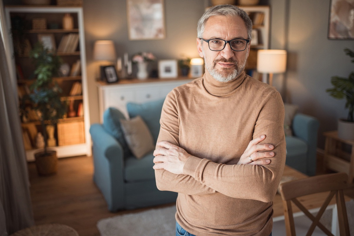 mężczyzna w wieku 50 plus w okularach stoi w salonie z zamyśloną miną