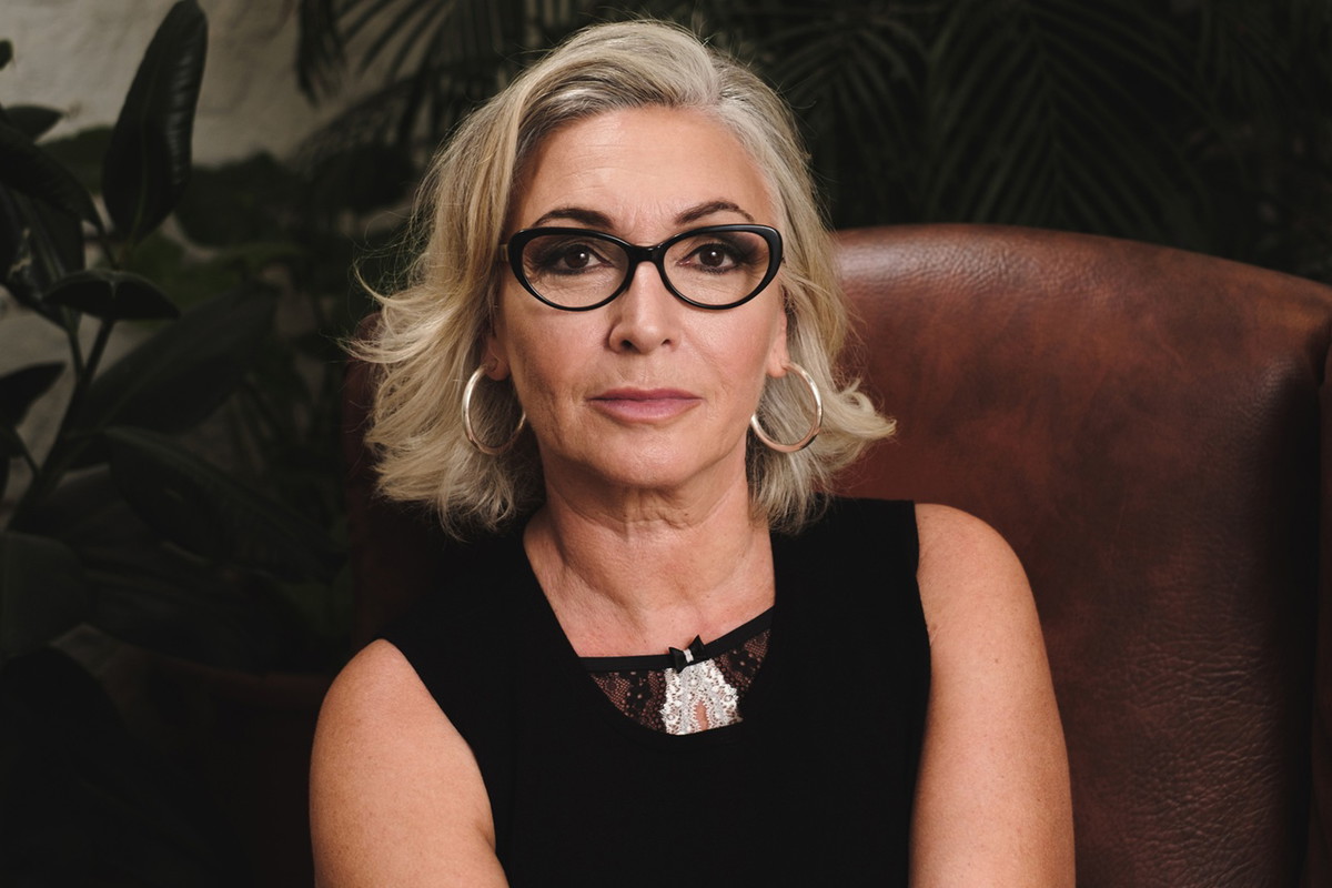 kobieta w w wieku 50 plus blondynka w okularach w czarnej sukience siedzi na skórzanym fotelu z poważną miną